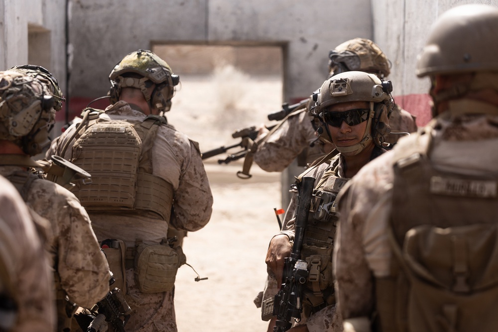 The image shows a group of military personnel in full combat gear conducting what appears to be a tactical training exercise or operation in an urban environment. The soldiers are wearing desert camouflage uniforms and are equipped with helmets, body armor, and firearms. They appear to be staging or moving through a breach point near a doorway in a damaged or austere structure.