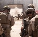 The image shows a group of military personnel in full combat gear conducting what appears to be a tactical training exercise or operation in an urban environment. The soldiers are wearing desert camouflage uniforms and are equipped with helmets, body armor, and firearms. They appear to be staging or moving through a breach point near a doorway in a damaged or austere structure.