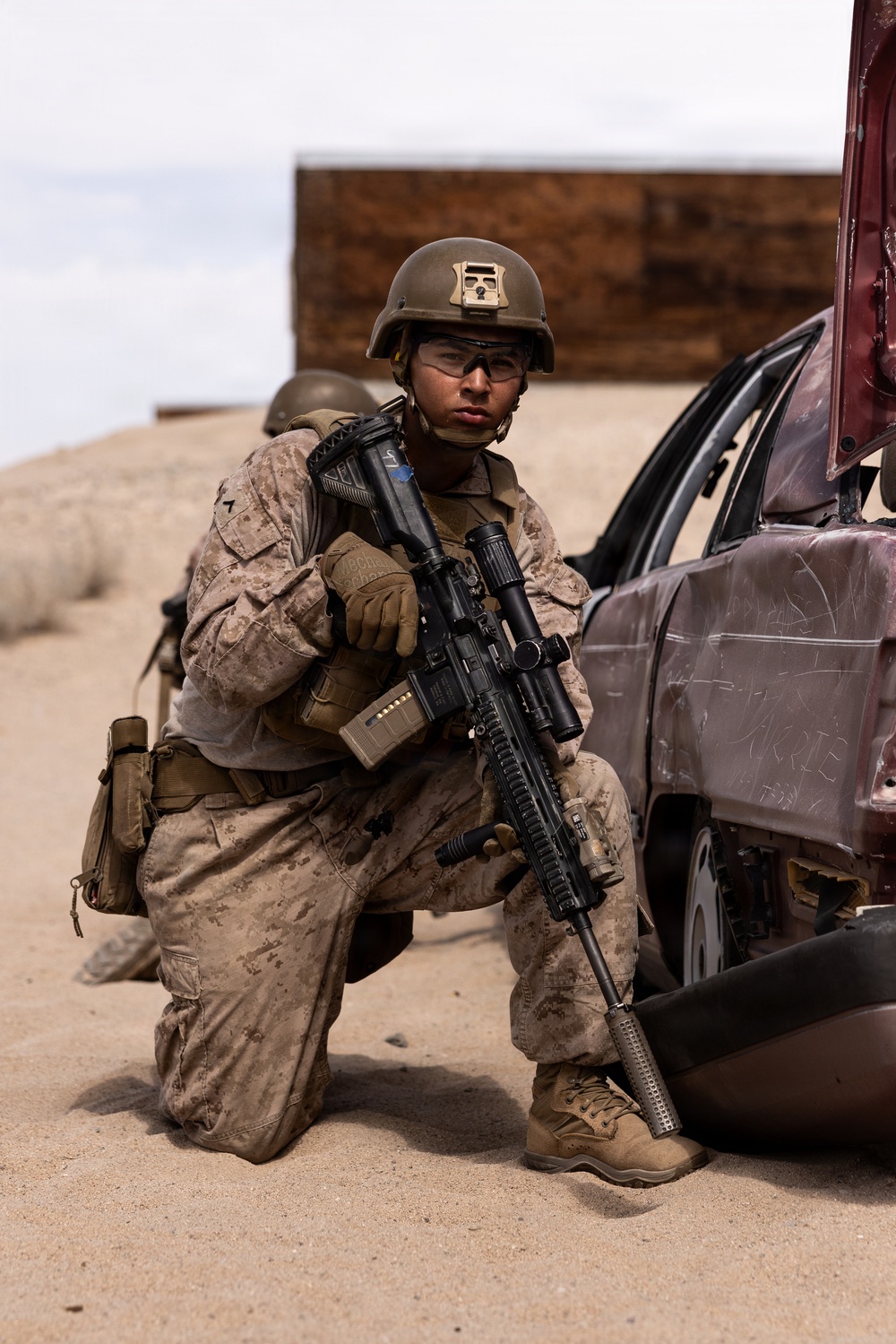 A U.S. Marine in full combat gear is kneeling beside a damaged vehicle in what appears to be a desert training environment. The Marine is equipped with a rifle featuring a scope, wearing MARPAT desert camouflage, a Kevlar helmet with a device mounted on top, and tactical gloves. A second helmeted figure is partially visible in the background.