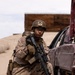A U.S. Marine in full combat gear is kneeling beside a damaged vehicle in what appears to be a desert training environment. The Marine is equipped with a rifle featuring a scope, wearing MARPAT desert camouflage, a Kevlar helmet with a device mounted on top, and tactical gloves. A second helmeted figure is partially visible in the background.