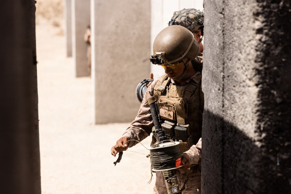 A military service member in desert camouflage uniform is handling a spool of wire or detonation cord while positioned against a concrete wall. The soldier is equipped with full tactical gear including a combat helmet with night vision mount, body armor, and yellow protective eyewear. The setting appears to be a training environment with multiple concrete block structures visible in the background.