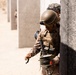 A military service member in desert camouflage uniform is handling a spool of wire or detonation cord while positioned against a concrete wall. The soldier is equipped with full tactical gear including a combat helmet with night vision mount, body armor, and yellow protective eyewear. The setting appears to be a training environment with multiple concrete block structures visible in the background.