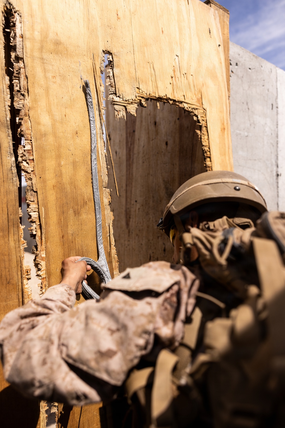 A military service member in desert camouflage (MARPAT pattern) and combat helmet is placing or handling what appears to be a breaching charge or det cord against a heavily damaged wooden door or wall panel. The wood shows significant splintering and blast damage consistent with prior breaching activity. This appears to be a training exercise at a military urban operations facility.