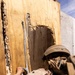 A military service member in desert camouflage (MARPAT pattern) and combat helmet is placing or handling what appears to be a breaching charge or det cord against a heavily damaged wooden door or wall panel. The wood shows significant splintering and blast damage consistent with prior breaching activity. This appears to be a training exercise at a military urban operations facility.