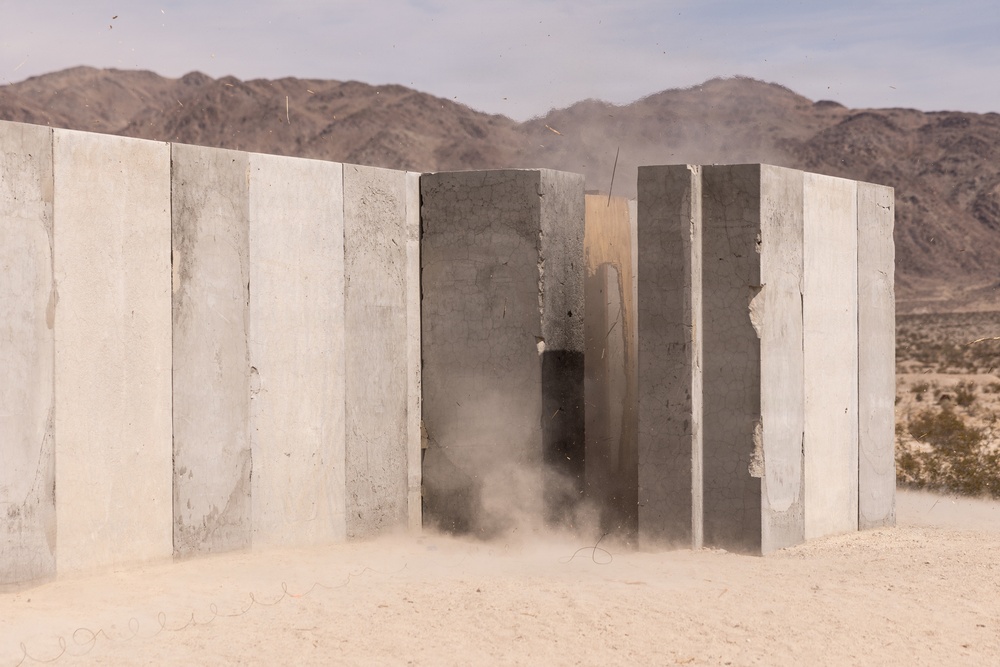 A concrete wall structure is being breached or demolished in a desert environment, with visible dust and debris indicating an active explosive or impact event. The wall consists of multiple vertical concrete panels, and a gap or breach is visible near the center where the disruption is occurring. The setting appears to be an arid desert landscape with mountains visible in the background.