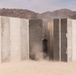 A concrete wall structure is being breached or demolished in a desert environment, with visible dust and debris indicating an active explosive or impact event. The wall consists of multiple vertical concrete panels, and a gap or breach is visible near the center where the disruption is occurring. The setting appears to be an arid desert landscape with mountains visible in the background.