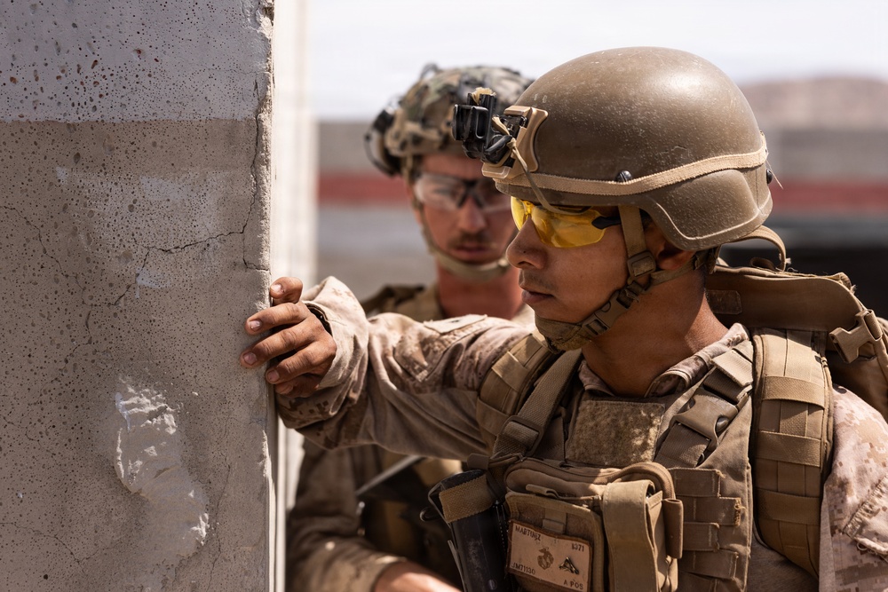 Two military personnel in desert camouflage uniforms and tactical gear are positioned against a concrete wall, appearing to conduct a tactical clearing or urban warfare exercise. The foreground soldier wears yellow-tinted ballistic eyewear, a tan combat helmet with night vision mount, and a plate carrier vest with visible name tape reading 'MARTUSZ'. The setting appears to be an arid or desert training environment.