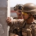 Two military personnel in desert camouflage uniforms and tactical gear are positioned against a concrete wall, appearing to conduct a tactical clearing or urban warfare exercise. The foreground soldier wears yellow-tinted ballistic eyewear, a tan combat helmet with night vision mount, and a plate carrier vest with visible name tape reading 'MARTUSZ'. The setting appears to be an arid or desert training environment.
