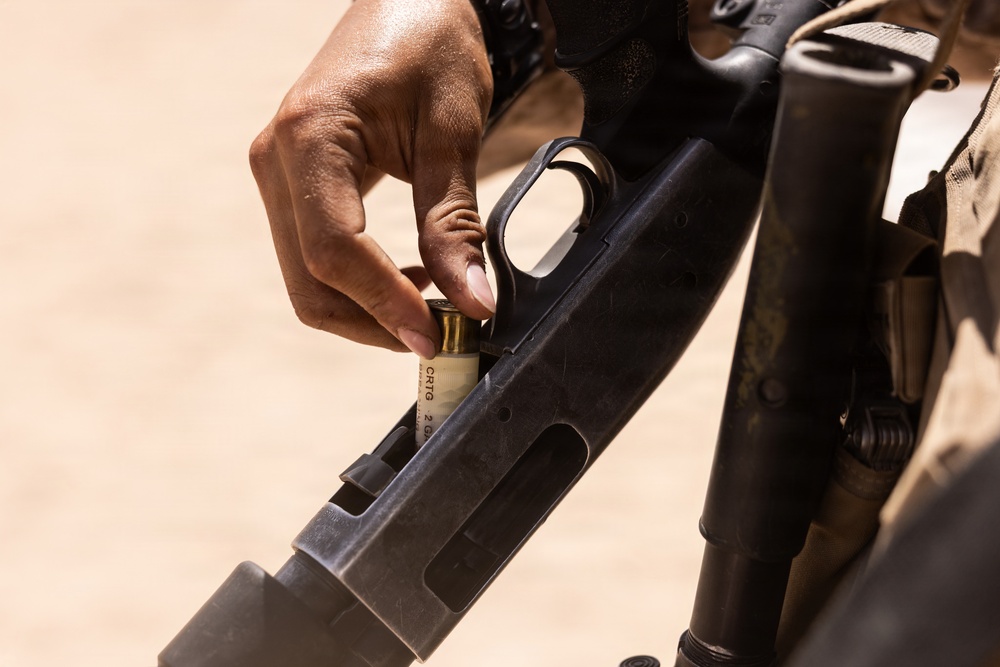 A close-up photograph shows a person's hand loading a shotgun shell into what appears to be a pump-action or semi-automatic shotgun. The shell is partially inserted into the loading port of the receiver. The individual appears to be wearing military or tactical gear based on visible clothing in the background.