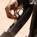 A close-up photograph shows a person's hand loading a shotgun shell into what appears to be a pump-action or semi-automatic shotgun. The shell is partially inserted into the loading port of the receiver. The individual appears to be wearing military or tactical gear based on visible clothing in the background.