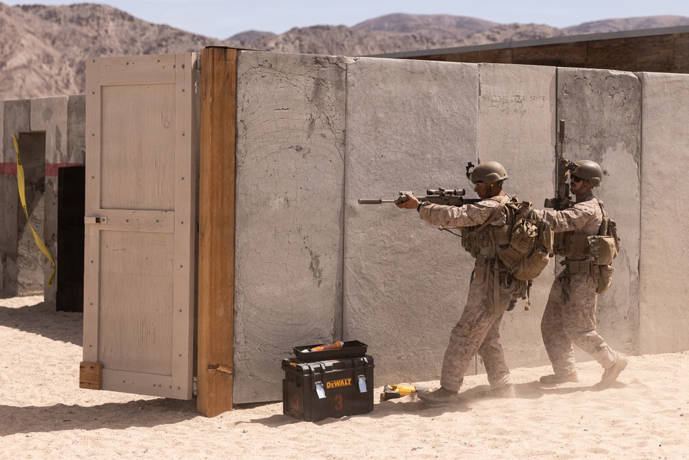 Two military personnel in desert camouflage uniforms are conducting a tactical training exercise at what appears to be a military training facility in an arid environment. They are moving in a stack formation around a concrete wall structure, with the lead soldier aiming a rifle equipped with a scope and suppressor. The setting includes simulated building structures, a wooden door frame, and a DeWalt tool case on the sandy ground.