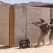 Two military personnel in desert camouflage uniforms are conducting a tactical training exercise at what appears to be a military training facility in an arid environment. They are moving in a stack formation around a concrete wall structure, with the lead soldier aiming a rifle equipped with a scope and suppressor. The setting includes simulated building structures, a wooden door frame, and a DeWalt tool case on the sandy ground.