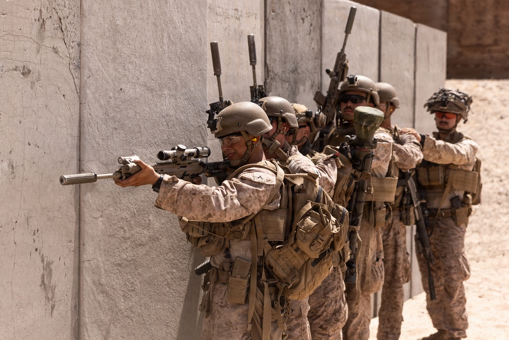 A line of military personnel in desert camouflage uniforms are positioned along a concrete wall in a tactical formation. The lead soldier is aiming a suppressed rifle with an optical scope, while others behind maintain a stacked formation. The setting appears to be a military training exercise or range environment based on the controlled conditions and sandy terrain.