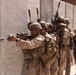 A line of military personnel in desert camouflage uniforms are positioned along a concrete wall in a tactical formation. The lead soldier is aiming a suppressed rifle with an optical scope, while others behind maintain a stacked formation. The setting appears to be a military training exercise or range environment based on the controlled conditions and sandy terrain.