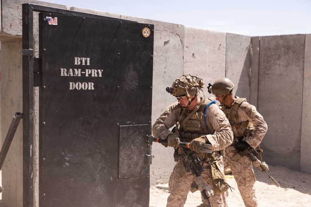 Two military personnel in desert camouflage uniforms are conducting breaching training on a BTI Ram-Pry Door at what appears to be a training facility. The soldiers are equipped with full tactical gear including helmets, body armor, and rifles. The setting features concrete barrier walls consistent with a military training range environment.