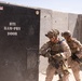 Two military personnel in desert camouflage uniforms are conducting breaching training on a BTI Ram-Pry Door at what appears to be a training facility. The soldiers are equipped with full tactical gear including helmets, body armor, and rifles. The setting features concrete barrier walls consistent with a military training range environment.