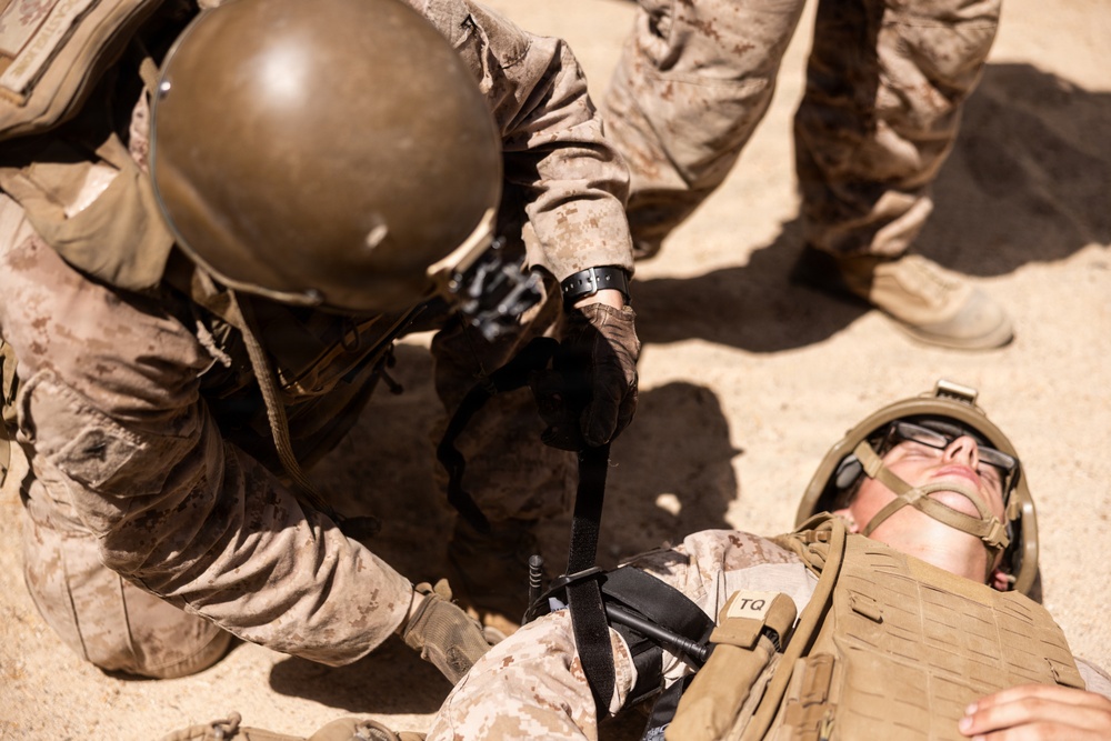The image shows military personnel in desert camouflage uniforms conducting what appears to be a combat casualty care drill or training exercise. One soldier is lying on the ground while another kneels over them applying what appears to be a tourniquet or medical device. The label 'TQ' visible on the downed soldier's gear is consistent with tourniquet marking, a standard tactical combat casualty care (TCCC) practice.