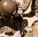 The image shows military personnel in desert camouflage uniforms conducting what appears to be a combat casualty care drill or training exercise. One soldier is lying on the ground while another kneels over them applying what appears to be a tourniquet or medical device. The label 'TQ' visible on the downed soldier's gear is consistent with tourniquet marking, a standard tactical combat casualty care (TCCC) practice.