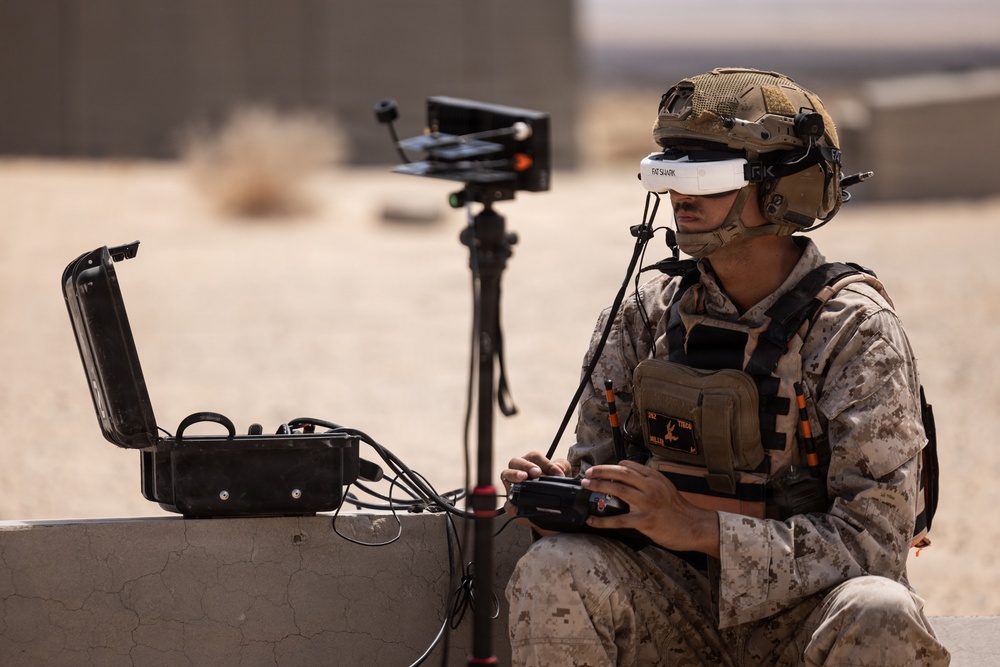 A military service member in desert camouflage uniform and tactical gear is operating a drone control system in an arid outdoor environment. The operator is wearing FatShark FPV (First Person View) goggles and holding a drone controller, with a ground station setup including a monitor on a tripod and a ruggedized equipment case. The setting appears to be a desert training or operational environment.