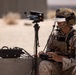 A military service member in desert camouflage uniform and tactical gear is operating a drone control system in an arid outdoor environment. The operator is wearing FatShark FPV (First Person View) goggles and holding a drone controller, with a ground station setup including a monitor on a tripod and a ruggedized equipment case. The setting appears to be a desert training or operational environment.