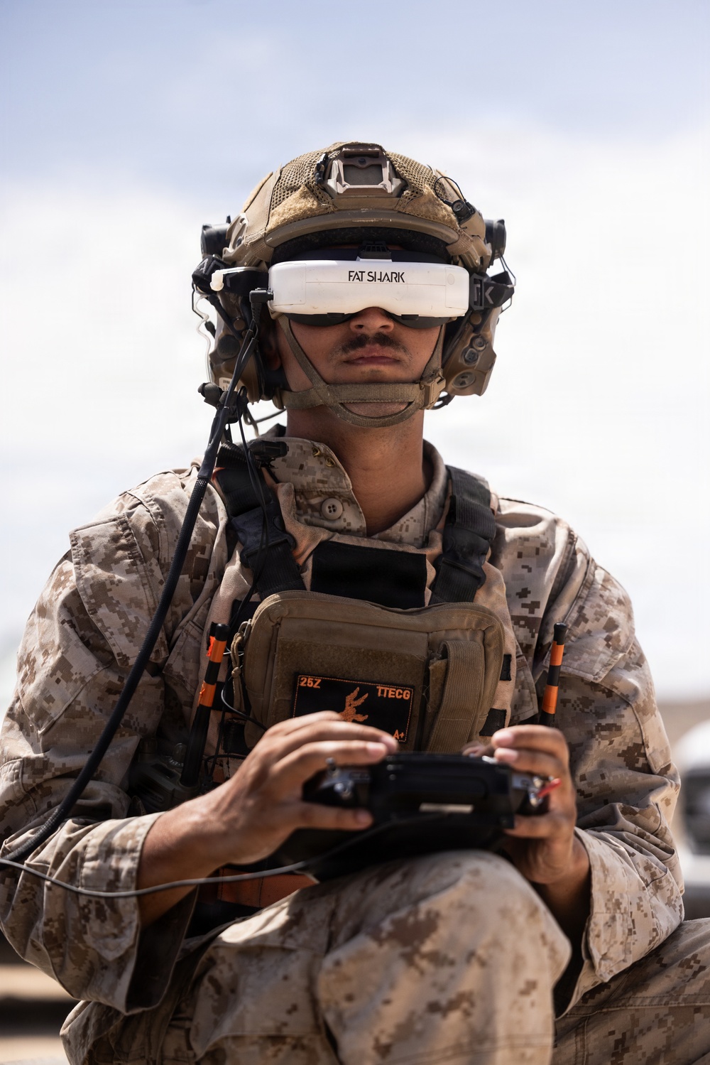 A military service member wearing USMC desert MARPAT camouflage uniform is operating a drone using FPV (First Person View) goggles branded 'Fat Shark' and a handheld remote controller. The individual is equipped with a tactical helmet, communications headset, body armor/plate carrier with visible patch markings '25Z', 'TTECG', and 'A+'. This appears to be a drone operator training or operational exercise.