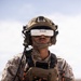 A military service member wearing USMC desert MARPAT camouflage uniform is operating a drone using FPV (First Person View) goggles branded 'Fat Shark' and a handheld remote controller. The individual is equipped with a tactical helmet, communications headset, body armor/plate carrier with visible patch markings '25Z', 'TTECG', and 'A+'. This appears to be a drone operator training or operational exercise.