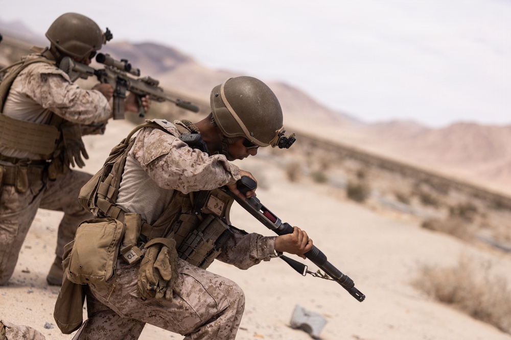 Two military personnel in desert camouflage uniforms are conducting what appears to be a training exercise in an arid desert environment. The foreground soldier is kneeling and aiming a shotgun-style weapon, while the background soldier is in a standing crouch aiming a scoped rifle. The setting, uniforms, and equipment are consistent with U.S. Marine Corps desert operations or training.