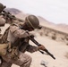 Two military personnel in desert camouflage uniforms are conducting what appears to be a training exercise in an arid desert environment. The foreground soldier is kneeling and aiming a shotgun-style weapon, while the background soldier is in a standing crouch aiming a scoped rifle. The setting, uniforms, and equipment are consistent with U.S. Marine Corps desert operations or training.