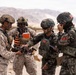 Four military personnel in different camouflage uniforms are gathered in a desert environment, examining a small drone or UAV. Two soldiers appear to be wearing US Marine Corps MARPAT desert camouflage, while two others wear a different digital camouflage pattern consistent with Republic of Korea (ROK) Marine Corps uniforms, suggesting a joint multinational training exercise. The arid, mountainous desert backdrop is consistent with locations such as the Mojave Desert in California.