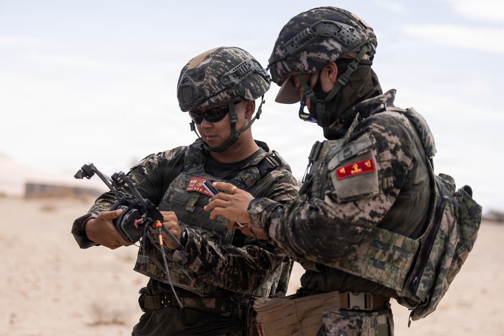 Two military personnel in camouflage uniforms are examining a small device, likely a drone or piece of tactical equipment, in an arid desert environment. Both soldiers are wearing tactical helmets and body armor with visible unit patches featuring Korean characters, suggesting they are members of the Republic of Korea (South Korean) armed forces. The setting and equipment suggest a training exercise or operational preparation.