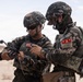 Two military personnel in camouflage uniforms are examining a small device, likely a drone or piece of tactical equipment, in an arid desert environment. Both soldiers are wearing tactical helmets and body armor with visible unit patches featuring Korean characters, suggesting they are members of the Republic of Korea (South Korean) armed forces. The setting and equipment suggest a training exercise or operational preparation.