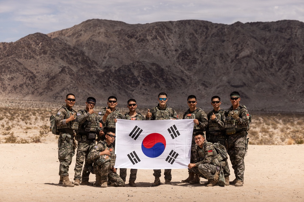 A group of approximately ten South Korean military personnel pose together in a desert environment holding the South Korean national flag (Taegukgi). The soldiers are dressed in camouflage combat uniforms and tactical gear, suggesting participation in a military exercise or training operation. The arid, mountainous landscape in the background is consistent with a desert training location.