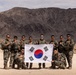 A group of approximately ten South Korean military personnel pose together in a desert environment holding the South Korean national flag (Taegukgi). The soldiers are dressed in camouflage combat uniforms and tactical gear, suggesting participation in a military exercise or training operation. The arid, mountainous landscape in the background is consistent with a desert training location.