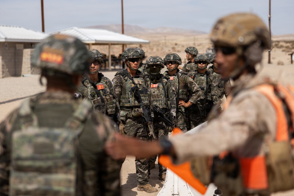 A group of military personnel in full combat gear are assembled in what appears to be a desert training environment. Soldiers wearing camouflage uniforms and tactical vests are being addressed or briefed by instructors, one of whom is wearing an orange safety vest. The setting suggests a joint military training exercise, likely at a desert training facility.