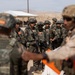 A group of military personnel in full combat gear are assembled in what appears to be a desert training environment. Soldiers wearing camouflage uniforms and tactical vests are being addressed or briefed by instructors, one of whom is wearing an orange safety vest. The setting suggests a joint military training exercise, likely at a desert training facility.
