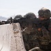 A soldier in Korean military camouflage uniform is positioned behind a concrete barrier, aiming a machine gun (appears to be an M60 or K3 light machine gun) in a desert training environment. Red and Korean-script patches are visible on the uniform, indicating Republic of Korea (ROK) Army affiliation. Additional soldiers are visible in the background in similar positions.