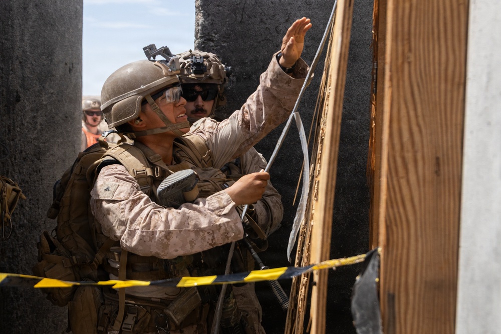 Two military personnel in desert camouflage uniforms are working with what appears to be a detonation cord or breaching cord attached to a wooden door or wall structure. A third individual in a hard hat and safety vest is visible in the background, suggesting this is a controlled training exercise. Yellow and black caution tape is visible in the foreground, reinforcing the controlled environment.