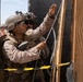 Two military personnel in desert camouflage uniforms are working with what appears to be a detonation cord or breaching cord attached to a wooden door or wall structure. A third individual in a hard hat and safety vest is visible in the background, suggesting this is a controlled training exercise. Yellow and black caution tape is visible in the foreground, reinforcing the controlled environment.