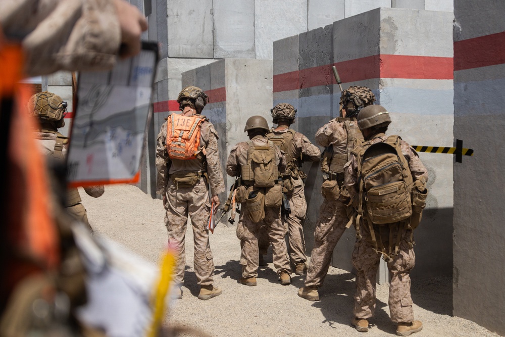 A group of military personnel in desert camouflage uniforms are gathered near a concrete barrier wall marked with red and white stripes. One individual wearing an orange backpack labeled '13E' appears to be in a leadership or observer role, while another person in the foreground is recording the activity on a tablet device. The setting appears to be a military training exercise at a constructed urban operations or breach training facility.