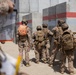 A group of military personnel in desert camouflage uniforms are gathered near a concrete barrier wall marked with red and white stripes. One individual wearing an orange backpack labeled '13E' appears to be in a leadership or observer role, while another person in the foreground is recording the activity on a tablet device. The setting appears to be a military training exercise at a constructed urban operations or breach training facility.