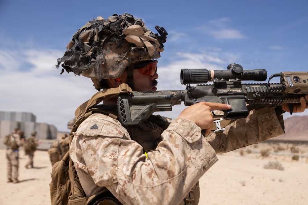 A U.S. Marine in desert MARPAT camouflage is shown in a combat training exercise in an arid desert environment. The Marine is aiming a rifle equipped with an optical scope while wearing a camouflaged helmet with netting and red-tinted protective eyewear. Additional personnel in similar uniforms are visible in the background.