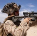 A U.S. Marine in desert MARPAT camouflage is shown in a combat training exercise in an arid desert environment. The Marine is aiming a rifle equipped with an optical scope while wearing a camouflaged helmet with netting and red-tinted protective eyewear. Additional personnel in similar uniforms are visible in the background.