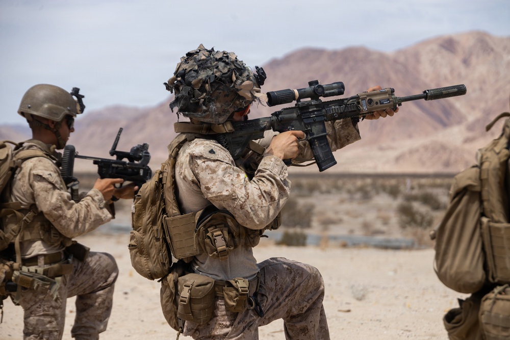 The image shows military personnel in desert camouflage uniforms conducting what appears to be a training exercise in an arid, desert environment. At least three soldiers are visible, equipped with tactical gear, body armor, and firearms. The setting features rocky desert terrain with reddish-brown mountains visible in the background.
