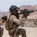 The image shows military personnel in desert camouflage uniforms conducting what appears to be a training exercise in an arid, desert environment. At least three soldiers are visible, equipped with tactical gear, body armor, and firearms. The setting features rocky desert terrain with reddish-brown mountains visible in the background.