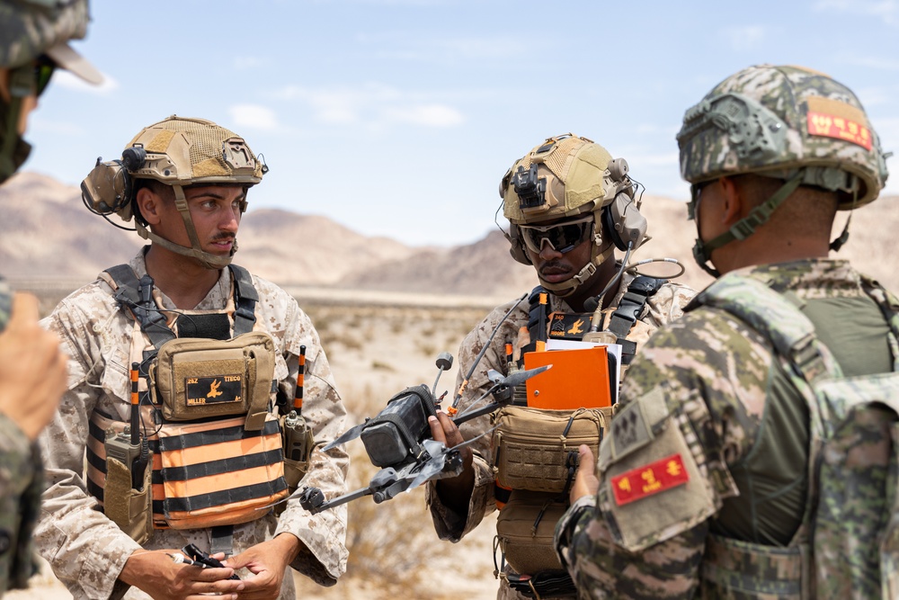Military personnel from what appear to be two different nations are gathered in a desert environment, examining a small drone. Two soldiers in US Marine Corps desert camouflage with orange training vests are interacting with a soldier in a different camouflage pattern bearing Korean-language insignia. The setting and equipment suggest a joint military training exercise.