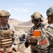 Military personnel from what appear to be two different nations are gathered in a desert environment, examining a small drone. Two soldiers in US Marine Corps desert camouflage with orange training vests are interacting with a soldier in a different camouflage pattern bearing Korean-language insignia. The setting and equipment suggest a joint military training exercise.
