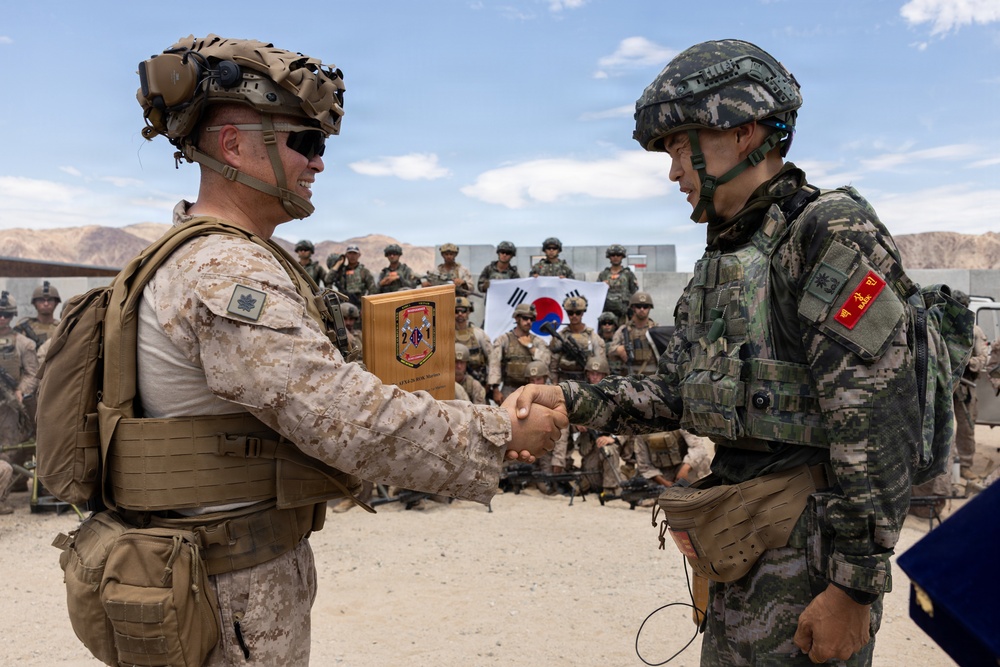 Two military personnel from different nations are shaking hands in what appears to be a joint military exercise or ceremony. One soldier wears U.S. Marine Corps desert MARPAT camouflage, while the other wears South Korean digital camouflage with Korean military patches. A wooden plaque is being exchanged, and a group of mixed military personnel stand in the background beneath a South Korean flag.