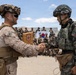 Two military personnel from different nations are shaking hands in what appears to be a joint military exercise or ceremony. One soldier wears U.S. Marine Corps desert MARPAT camouflage, while the other wears South Korean digital camouflage with Korean military patches. A wooden plaque is being exchanged, and a group of mixed military personnel stand in the background beneath a South Korean flag.