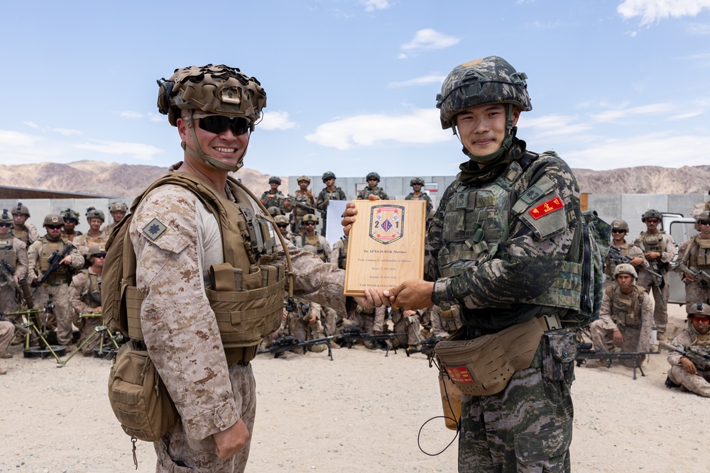 Two military personnel — one in US Marine Corps desert camouflage and one in Republic of Korea (ROK) Marine Corps digital camouflage — are shaking hands while jointly holding a wooden commemorative plaque. The plaque bears a unit insignia with the number '2/1' and text referencing 'AFSA-26 ROK Marines.' A large group of soldiers from both nations is assembled in the background at what appears to be a desert training facility.