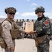 Two military personnel — one in US Marine Corps desert camouflage and one in Republic of Korea (ROK) Marine Corps digital camouflage — are shaking hands while jointly holding a wooden commemorative plaque. The plaque bears a unit insignia with the number '2/1' and text referencing 'AFSA-26 ROK Marines.' A large group of soldiers from both nations is assembled in the background at what appears to be a desert training facility.